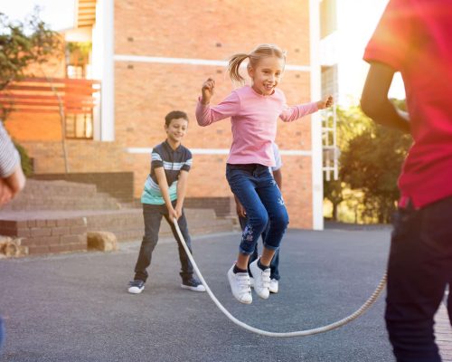 Happy elementary kids playing together with jumping rope outdoor. Children playing skipping rope jumping game and laughing outdoors. Happy cute girl jumping over skipping rope held by her friends.