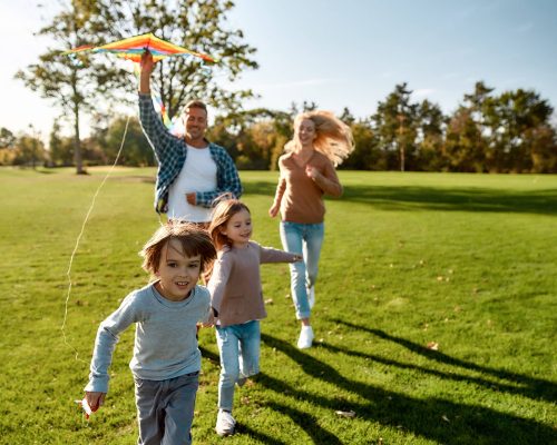 Portrait of cheerful parents with two kids running with kite in the park on a sunny day. Family, kids and nature concept. Horizontal shot.