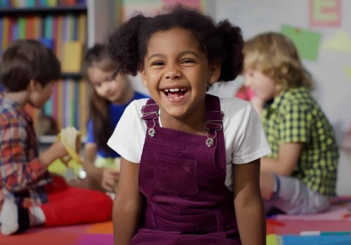 Close up portrait of smiling little African-American girl looking at camera at primary school. Adorable preschool kid sitting in playroom of kindergarten